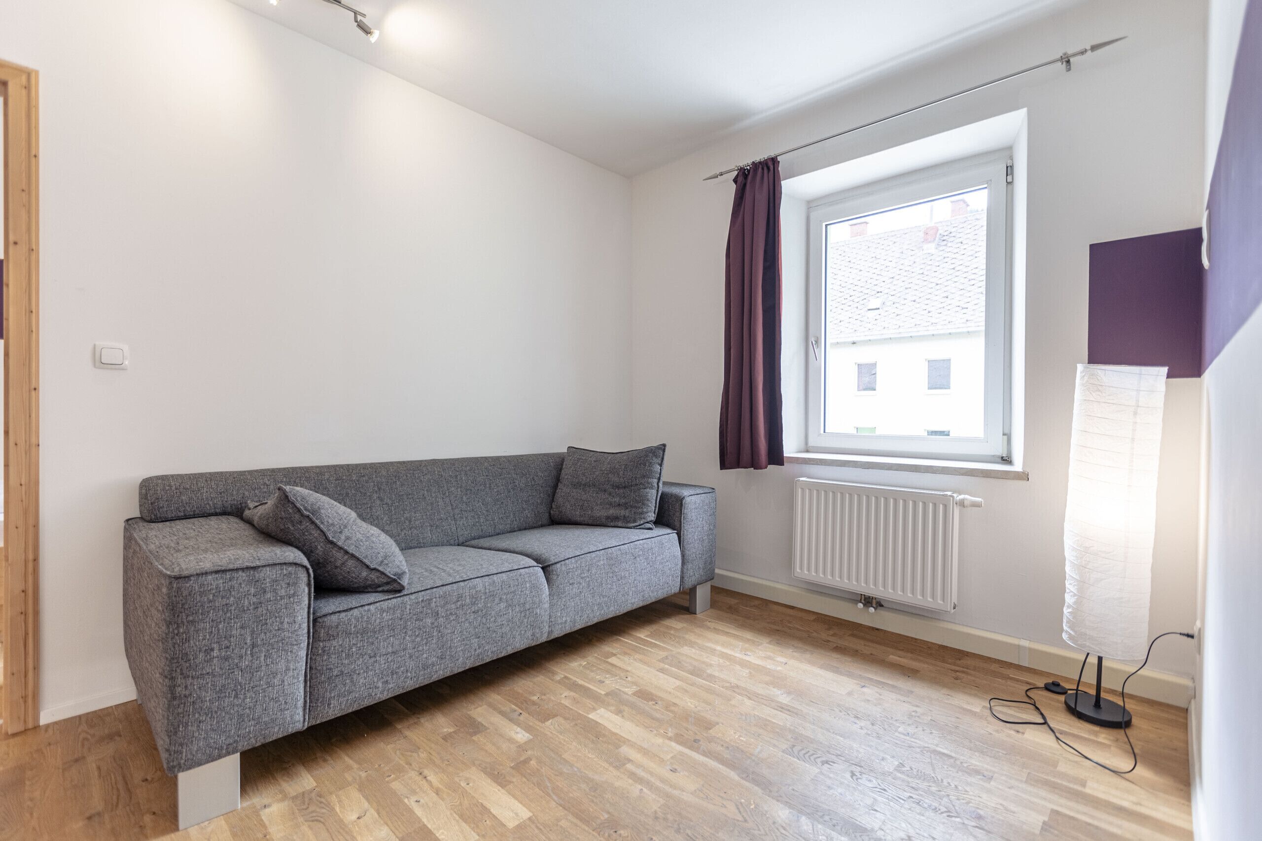 A modern, sparsely furnished room with a grey sofa, a window with curtains, and a radiator.