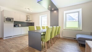 Une cuisine moderne dotée d’un four à micro-ondes encastré, un coin repas avec une table grise et des chaises vertes, et une fenêtre offrant une vue sur une montagne.