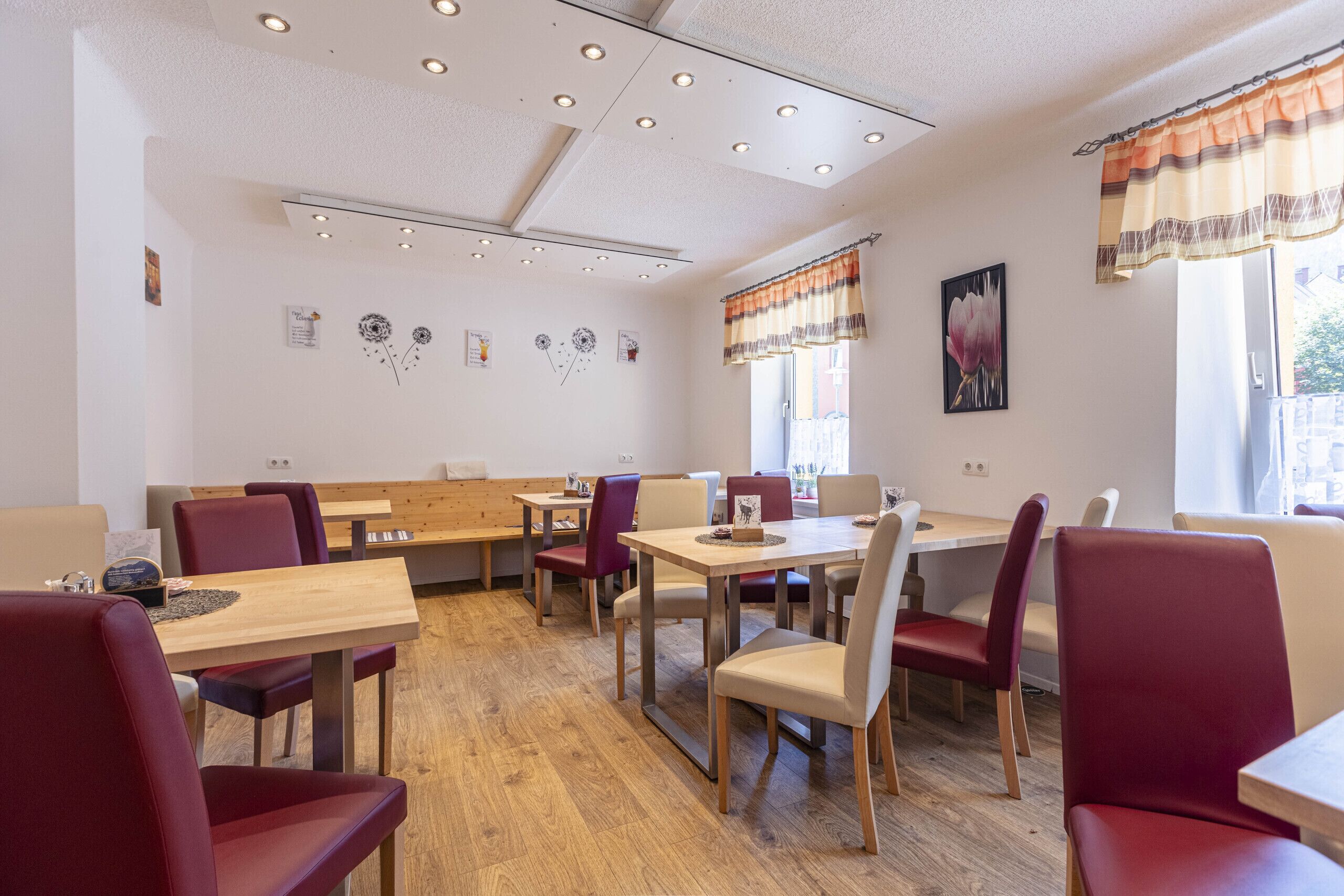 A dining area with wooden tables and red chairs, a ceiling with recessed lighting, and artwork on the walls.