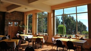 Un espace repas avec des tables et des chaises, de grandes fenêtres offrant une vue sur les arbres et les montagnes, et un mur en pierre.