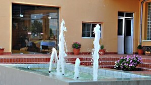 Une fontaine avec des jets d’eau se trouve devant un bâtiment dont l’entrée est recouverte de carreaux et qui est agrémenté de plantes en pot.