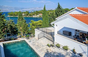 A poolside area with a stone deck, a white building with a red-tiled roof, and a view of the sea and mountains in the background.
