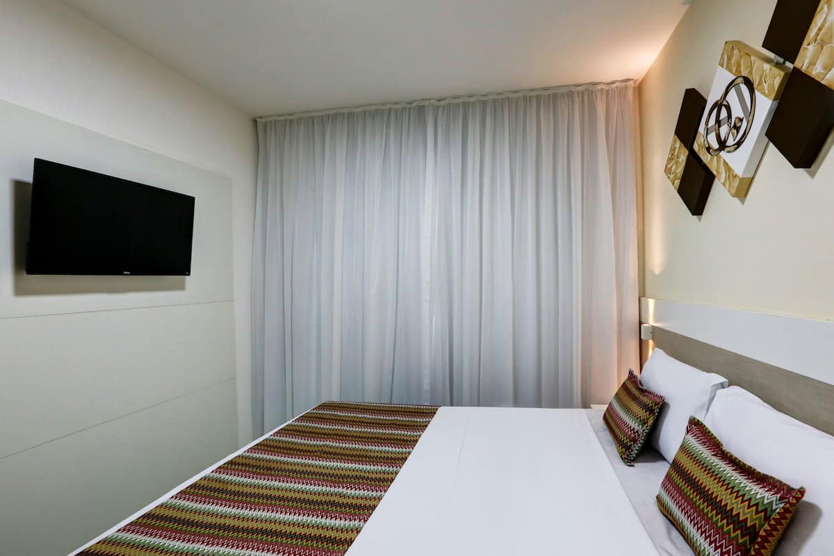 A hotel room with a bed, a flat-screen TV mounted on the wall, a large window with white curtains, and a wall-mounted artwork.