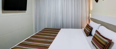A hotel room with a bed, a flat-screen TV mounted on the wall, a large window with white curtains, and a wall-mounted artwork.