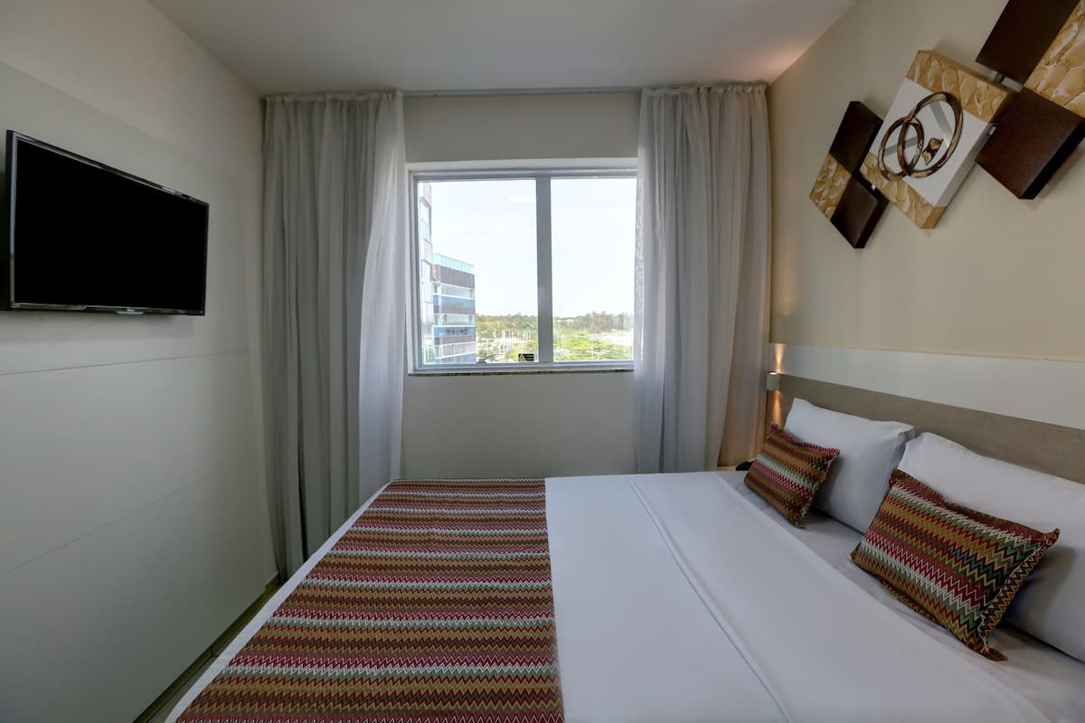 A hotel room with a bed, a TV mounted on the wall, a window with curtains, and a view of buildings outside.