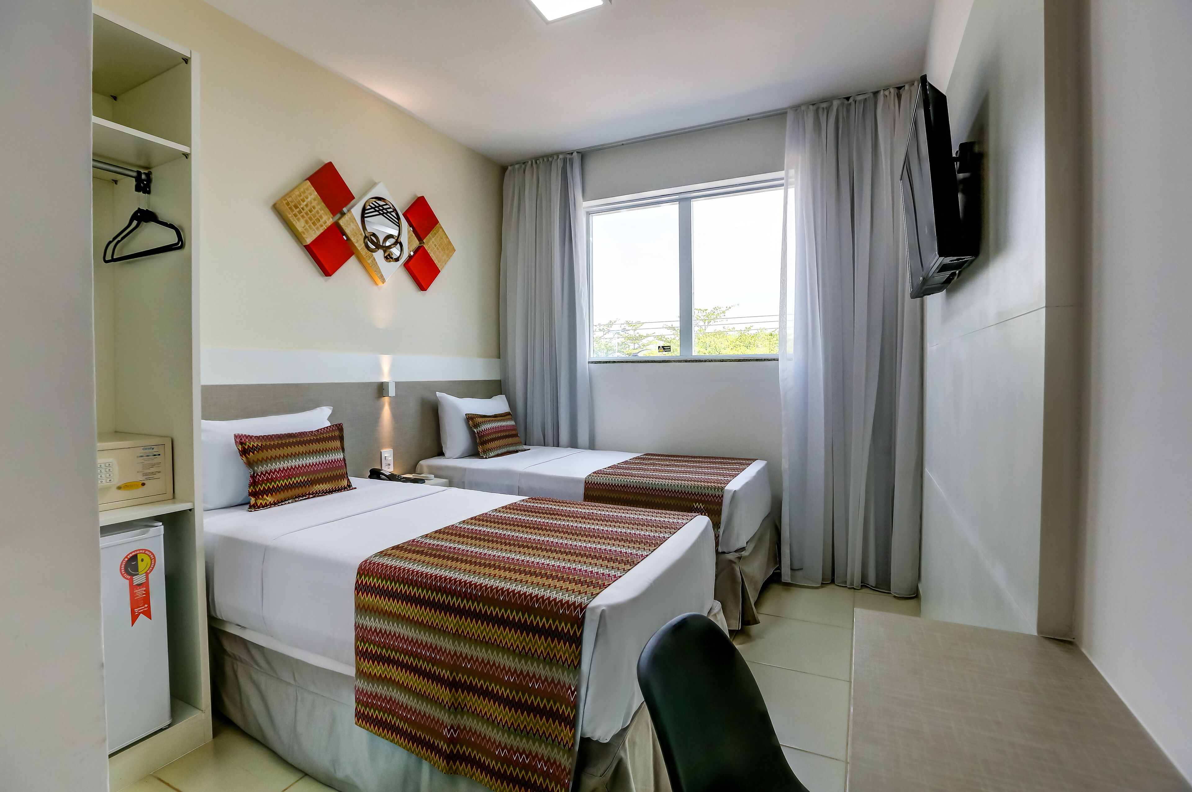 A hotel room with two beds, a TV, a desk, a chair, a window with curtains, and a wall decoration.