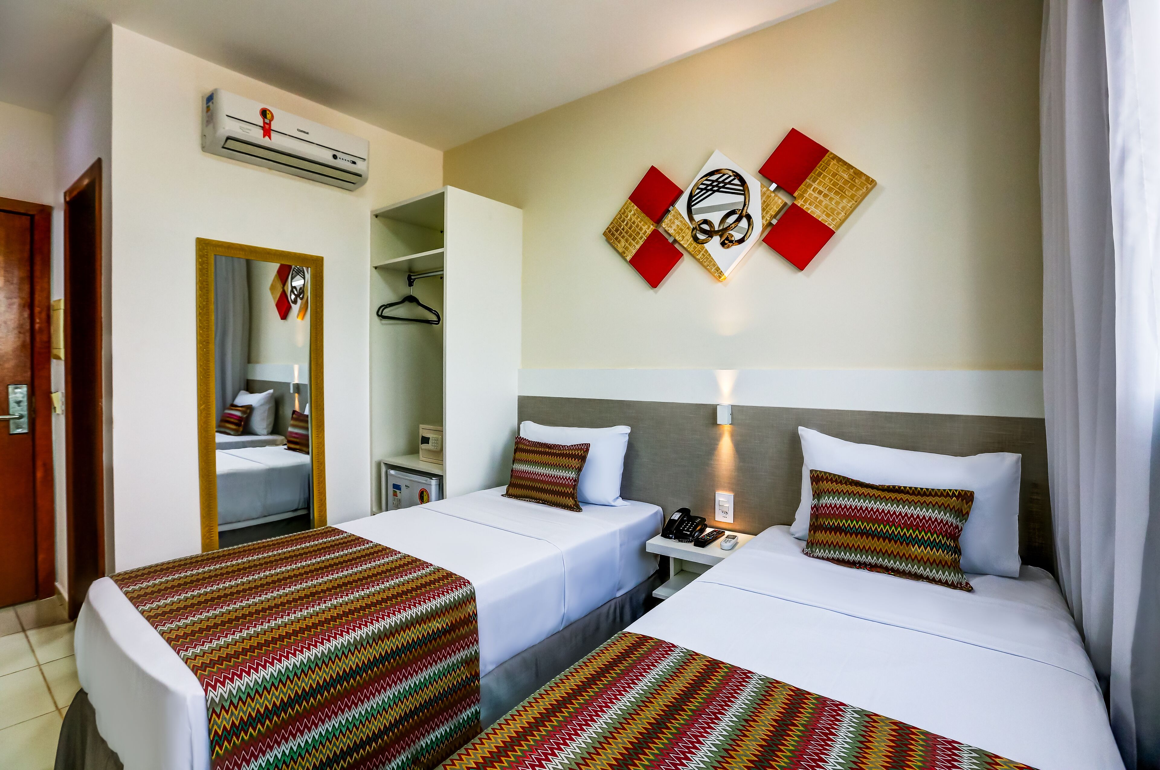 A hotel room with two beds, a mirror, a wardrobe, and a wall-mounted air conditioner.