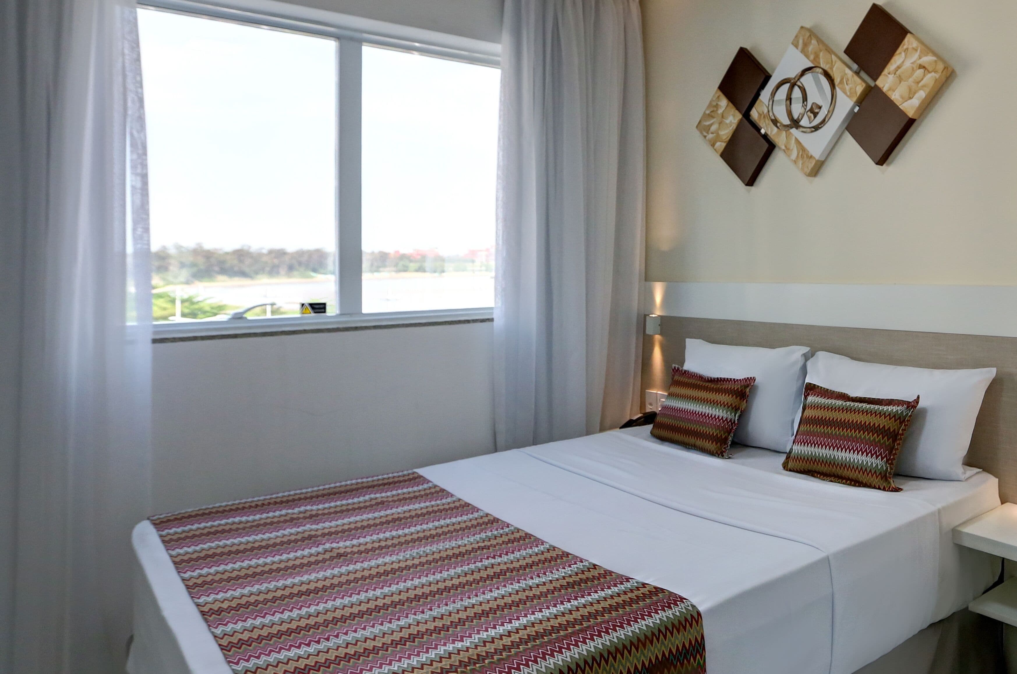 Hotellihuone, jossa on suuri ikkuna, valkoisilla lakanoiden peitetty sänky, kaksi koristeellista tyynyä ja seinälle ripustettu taideteos.