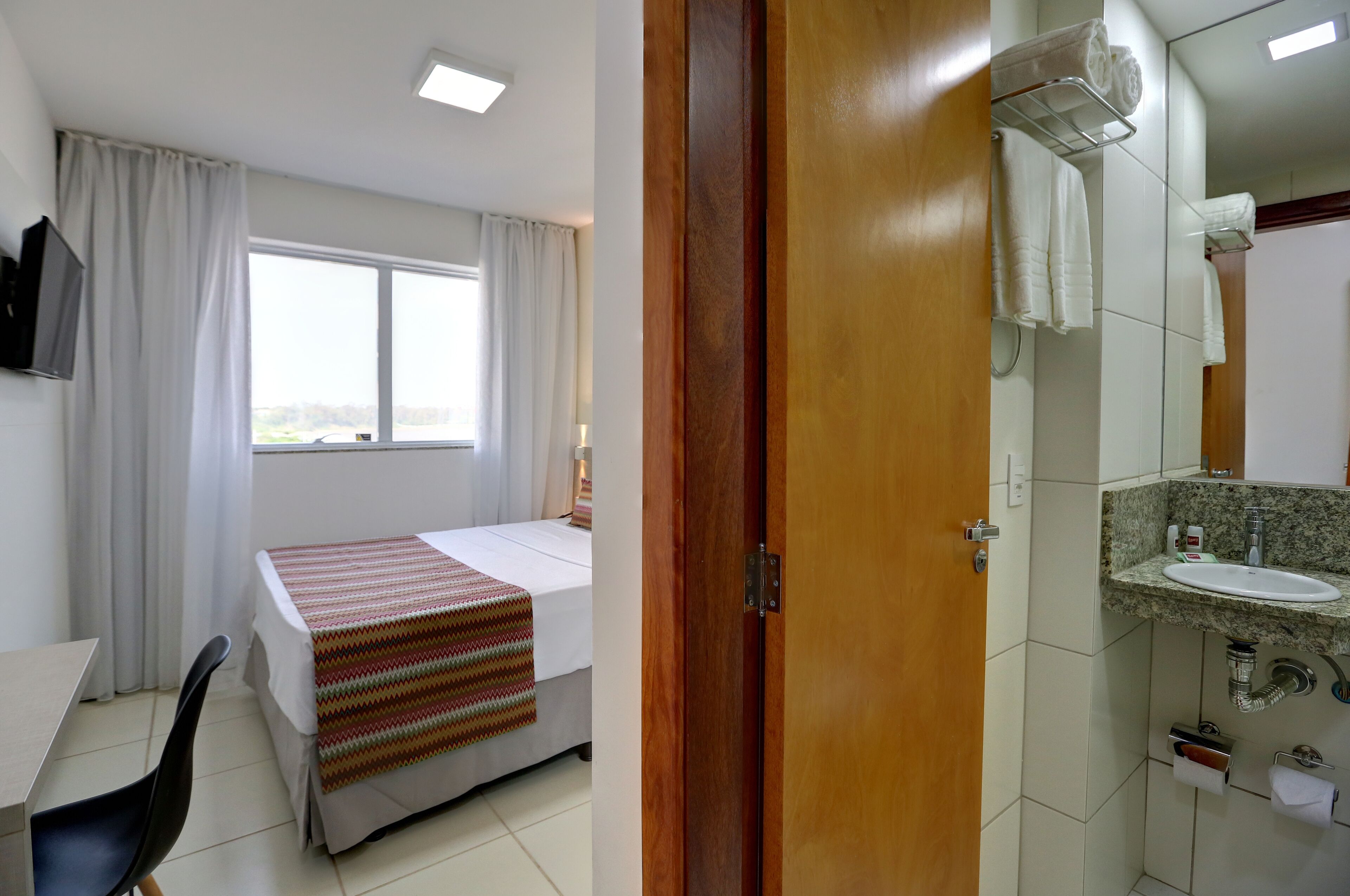 A hotel room with a bed, a desk, a chair, a window with curtains, a wooden door, and a bathroom with a sink and mirror.