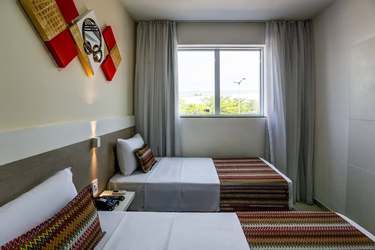 A hotel room with two beds, a window with curtains, a wall-mounted light, and a view of greenery outside.