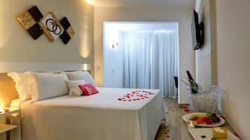 A hotel room with a neatly made bed, a bedside table with a bottle and glasses, and a fruit plate on the floor.