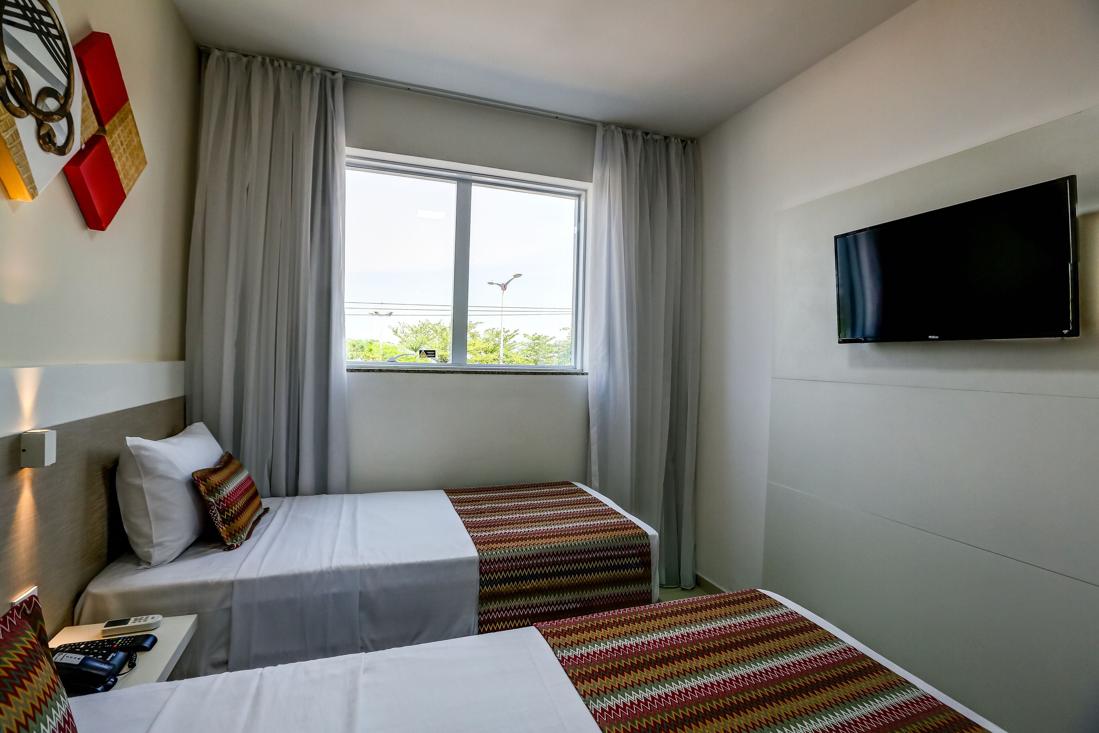 A hotel room with two beds, a TV, a window with curtains, and a view of greenery outside.