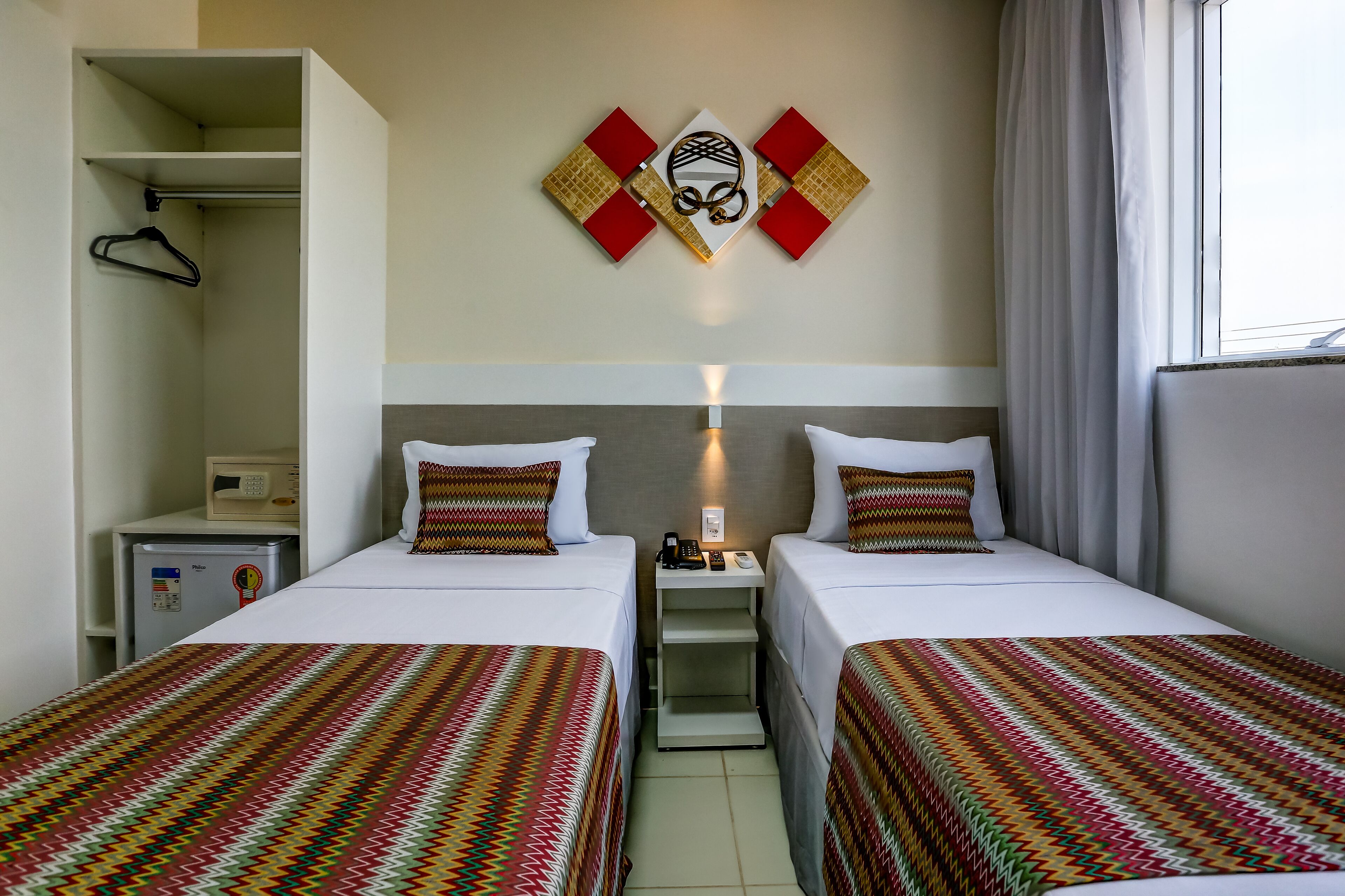 Habitación de hotel con dos camas, una mesita de noche con teléfono y lámpara, un escritorio pequeño con ordenador y una obra de arte colgada en la pared.