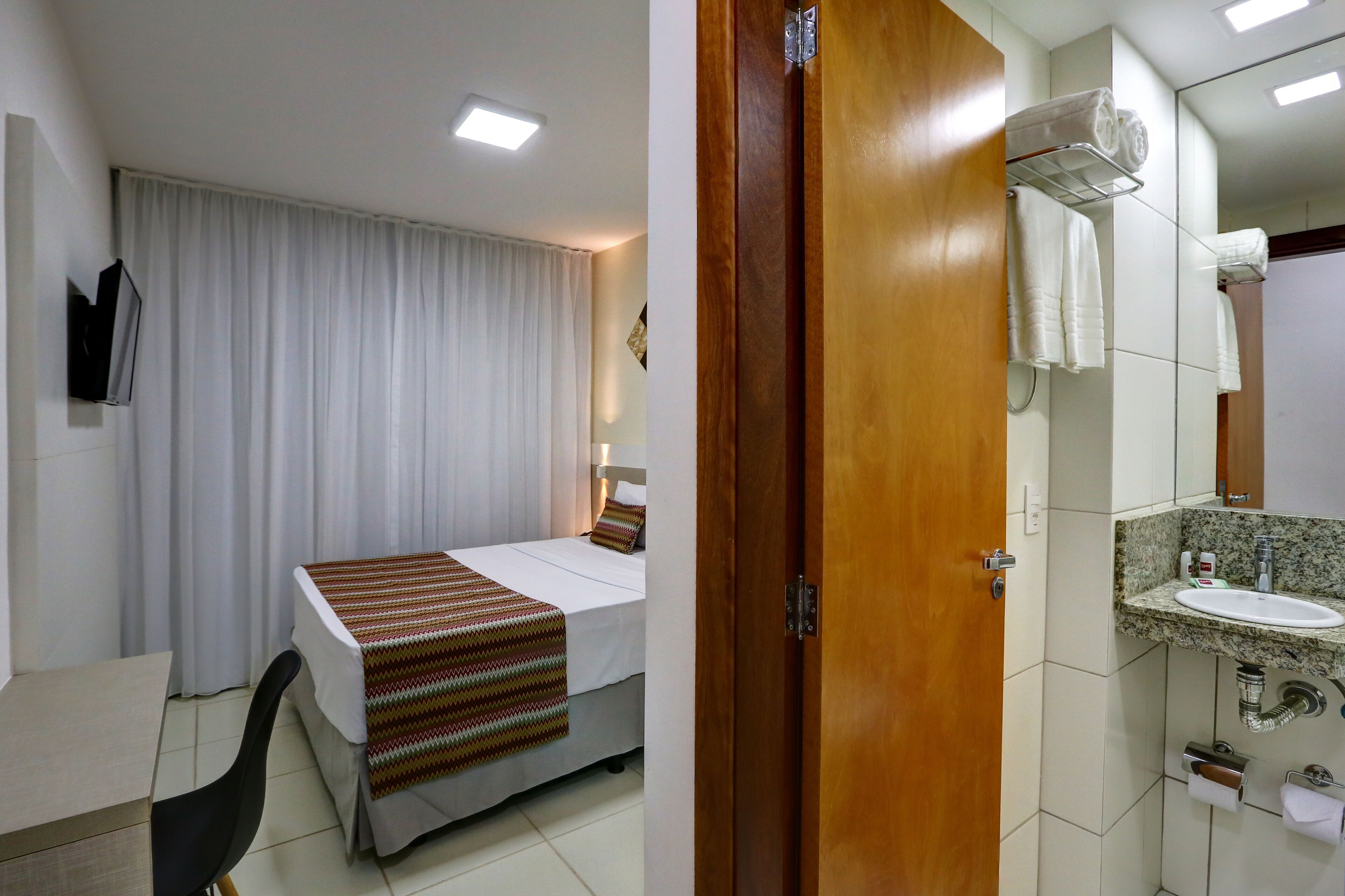 A hotel room with a bed, a desk, a chair, a bathroom with a sink and mirror, and a shower.