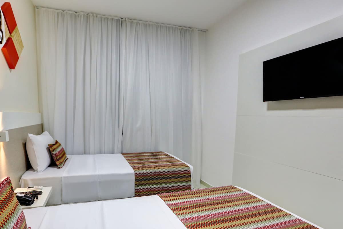 A hotel room with two beds, a TV mounted on the wall, and a large window with curtains.