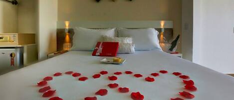 A hotel room with a neatly made bed, a heart-shaped arrangement of rose petals, a red box, and a small plate with a candle.
