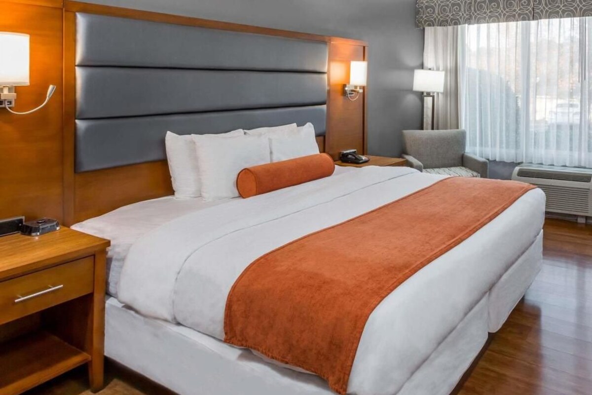 A hotel room with a large bed, a bedside table, a nightstand, a lamp, a chair, and a window with curtains.