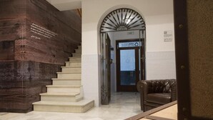 Una escalera con una pared oscura de madera a un lado y un piso de baldosas blancas. Hay un sofá de cuero marrón y una puerta con un letrero encima.