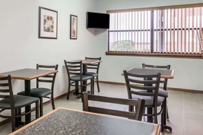A dining area with tables and chairs, a flat-screen TV mounted on the wall, and a window with vertical blinds.
