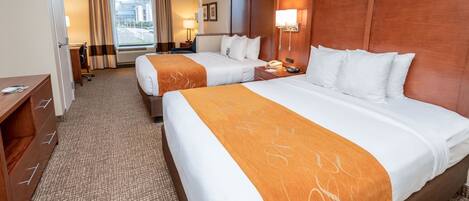 A hotel room with two beds, a desk, and a view of the city through large windows.