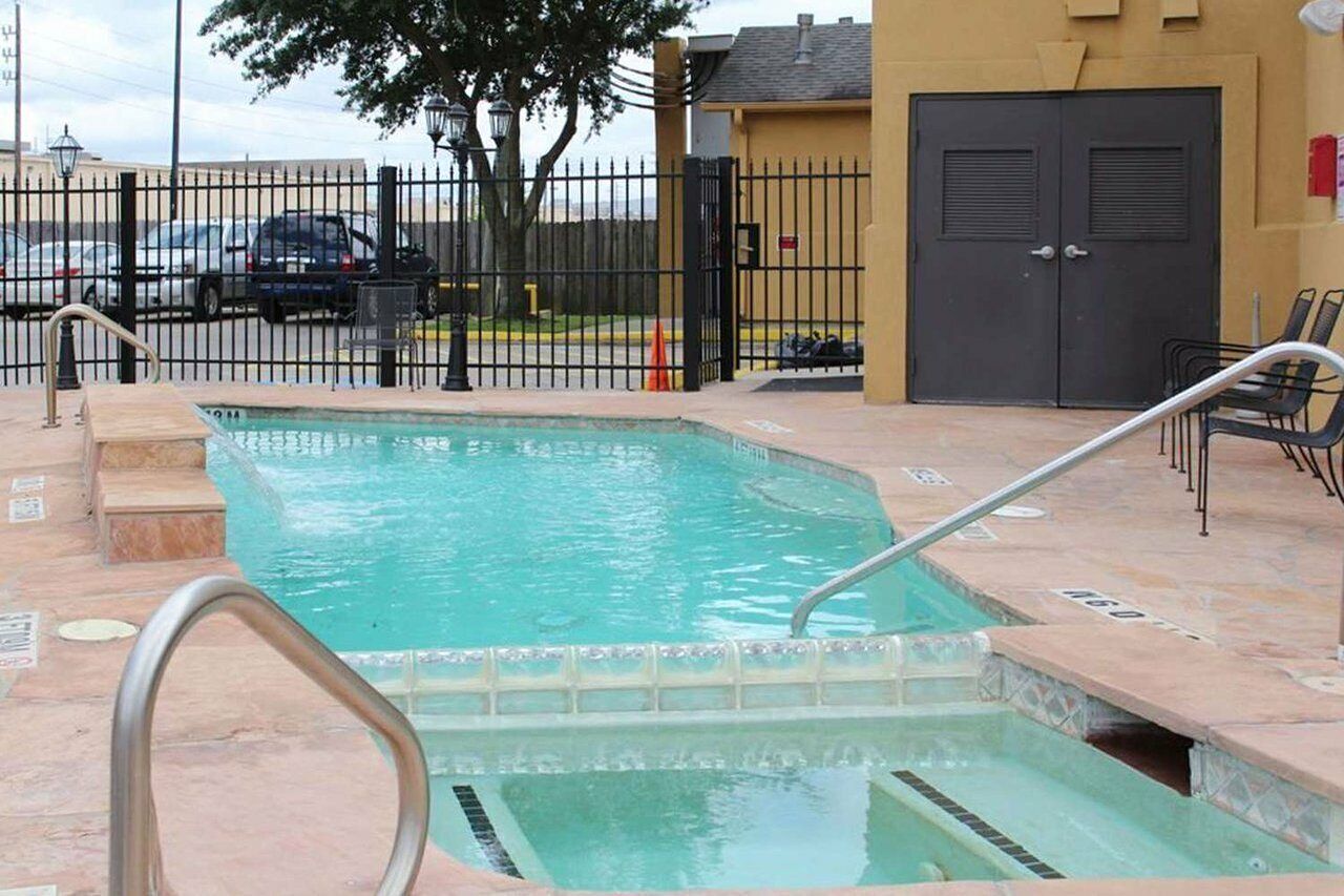 Una piscina rectangular con una escalera metálica y escalones, rodeada por una valla y un edificio con una puerta.