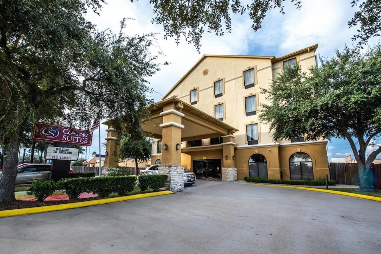 Entrada de hotel con un letrero de Comfort Suites, zona de estacionamiento techada y cielo despejado.