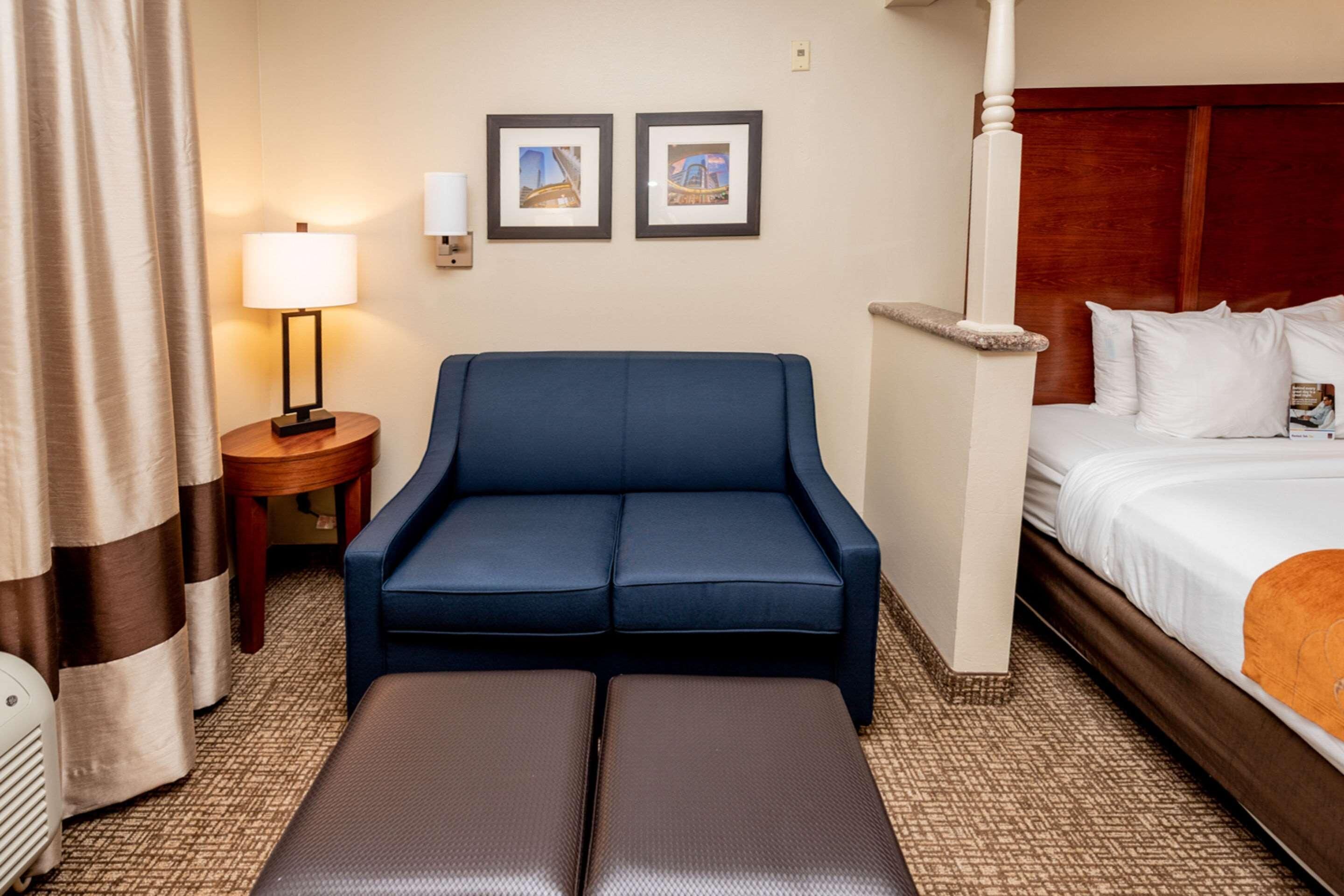 Een hotelkamer met een blauwe bank, een bed met witte lakens, een klein tafeltje met een lamp en twee ingelijste foto's aan de muur.