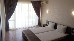 A hotel room with two beds, a window with curtains, an air conditioning unit, and a wall-mounted light. - Marack2 (Nessebar)
