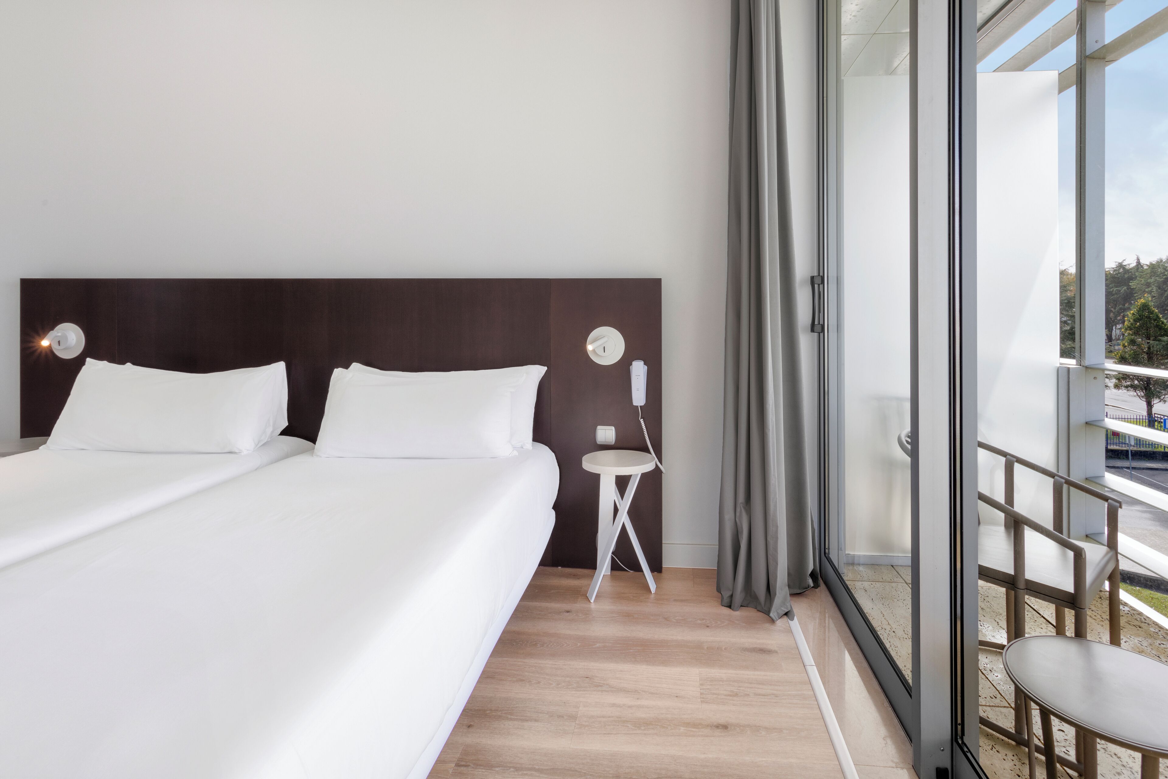 Ein modernes Hotelzimmer mit einem großen Bett, einem kleinen Nachttisch und Blick durch eine Glastür.