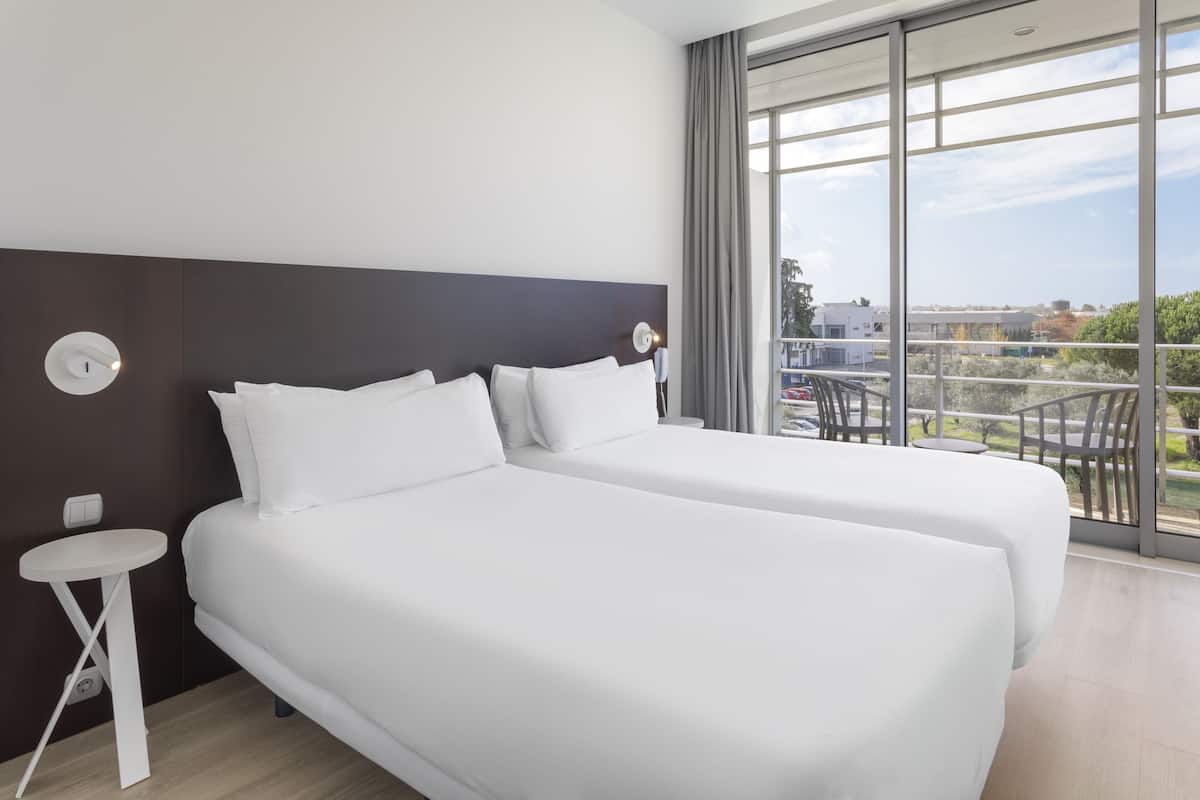 Une chambre d’hôtel avec deux lits, une petite table blanche, une grande fenêtre donnant sur la ville et un balcon avec des chaises.