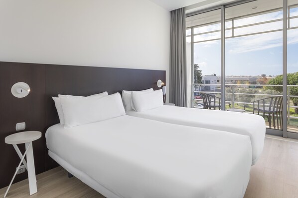 Habitación de hotel con dos camas, una mesita pequeña blanca, un ventanal con vistas a la ciudad y un balcón con sillas.