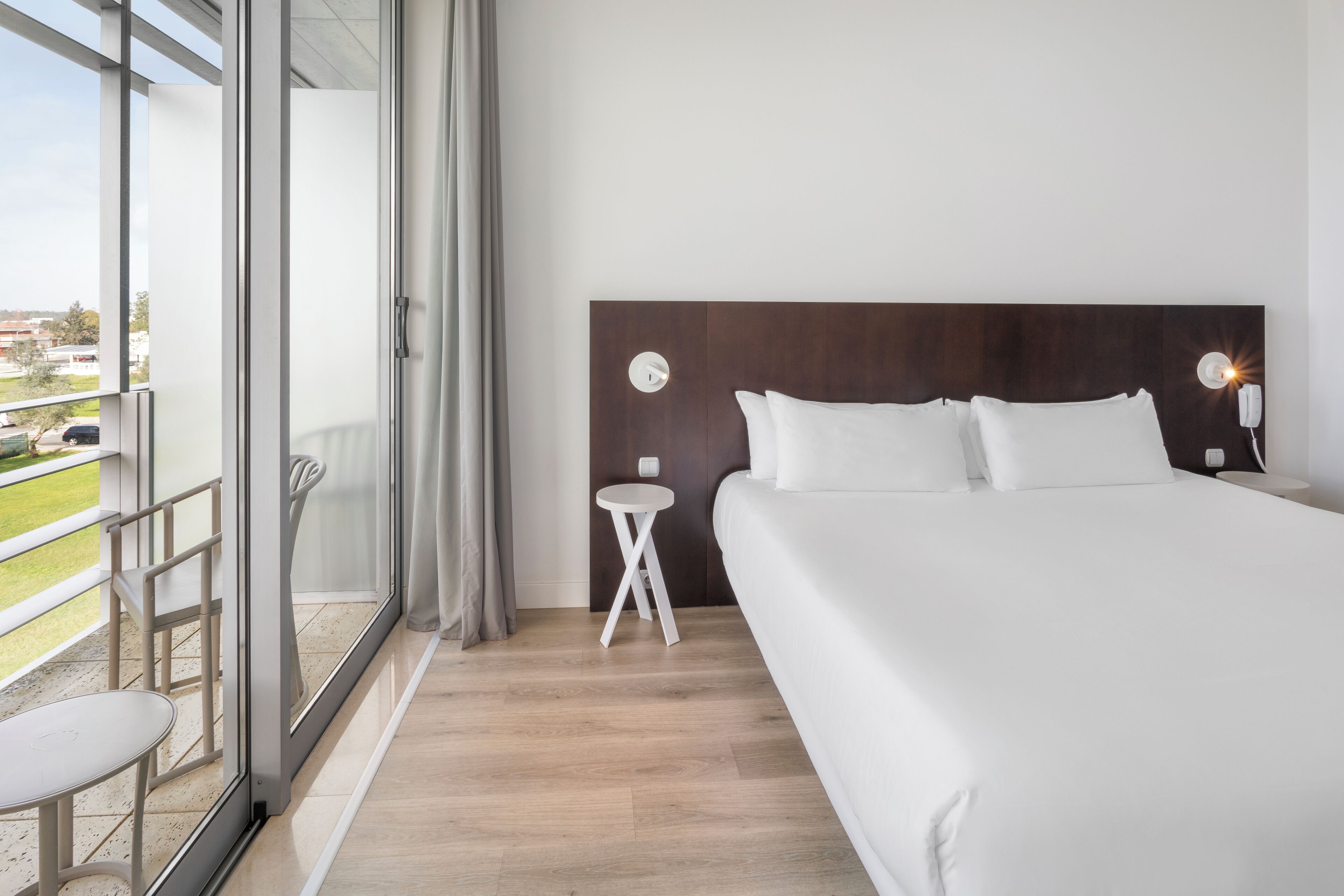 Hotellihuone, jossa on suuri sänky, pieni sivupöytä, yöpöytä lampun kanssa ja liukuovista lasiterassia vastapäätä avautuva näkymä ulos.