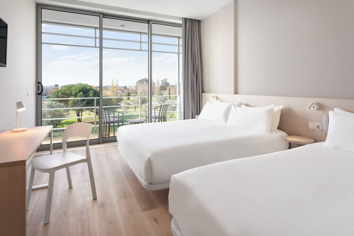 Une chambre d’hôtel avec deux lits, un bureau, une chaise et une grande fenêtre donnant sur l’extérieur.