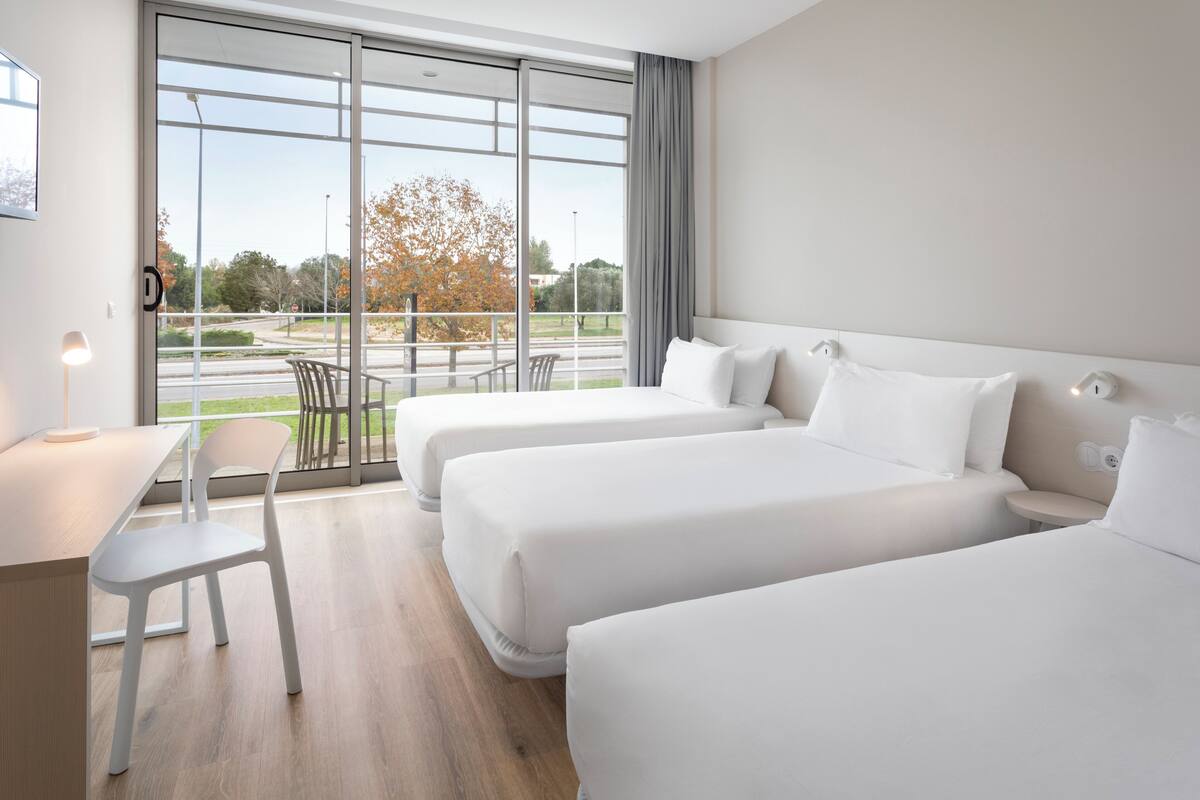 Une chambre d’hôtel avec deux lits, un bureau, une chaise et une grande fenêtre donnant sur l’extérieur.