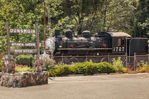Una locomotora de vapor antigua con el número 1727 en una estación de tren, rodeada de follaje y un letrero que indica 'DUNSMUIR BOTANICAL GARDENS RIVER ACCESS'.