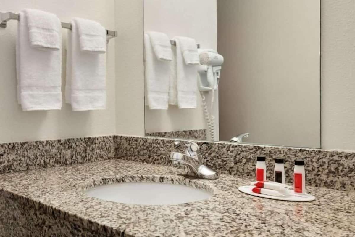 Un baño con encimera de granito, un lavabo, un espejo, un perchero con toallas, un secador de pelo y artículos de aseo.