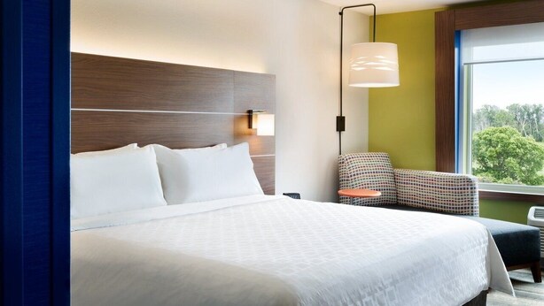 A hotel room with a large bed, a headboard, a wall-mounted lamp, a chair, and a window with a view. - Holiday Inn Express Canton Potsdam (Canton)