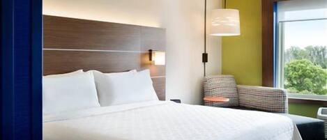 A hotel room with a large bed, a nightstand, a lamp, and a view of the outdoors.