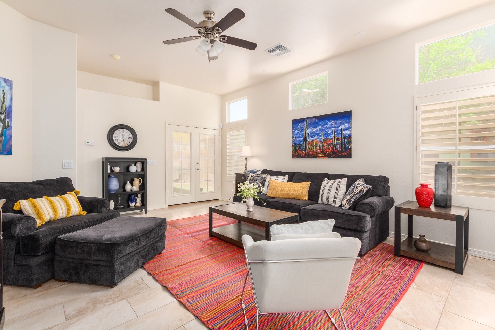 A spacious living room with a dark grey sofa, a white armchair, and a coffee table. There is a colorful rug on the floor, a ceiling fan, and a painting on the wall.