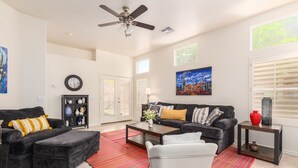 A spacious living room with a dark grey sofa, a white armchair, and a coffee table. There is a colorful rug on the floor, a ceiling fan, and a painting on the wall. - Ahwatukee/Foothills-Golfer's Paradise! Desert Vacation Retreat/Gated Community! (Phoenix)