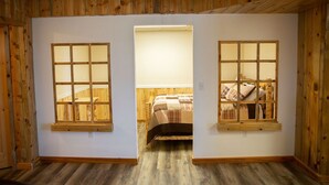 A room with wooden walls and floors, a bed with a plaid bedspread, and two windows with wooden frames.