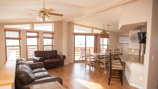 A spacious living area with a leather sofa, wooden coffee table, and a dining table with chairs. There is a ceiling fan, large windows, and a kitchen area with a clock and a TV.