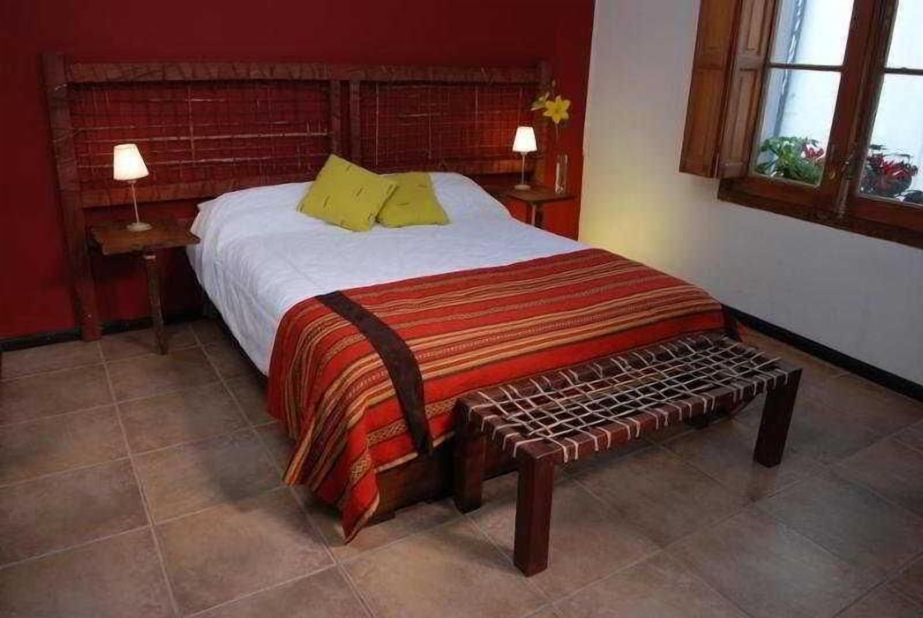 A bed with a wooden headboard, two bedside lamps, a bench at the foot of the bed, and a window with a view of plants.