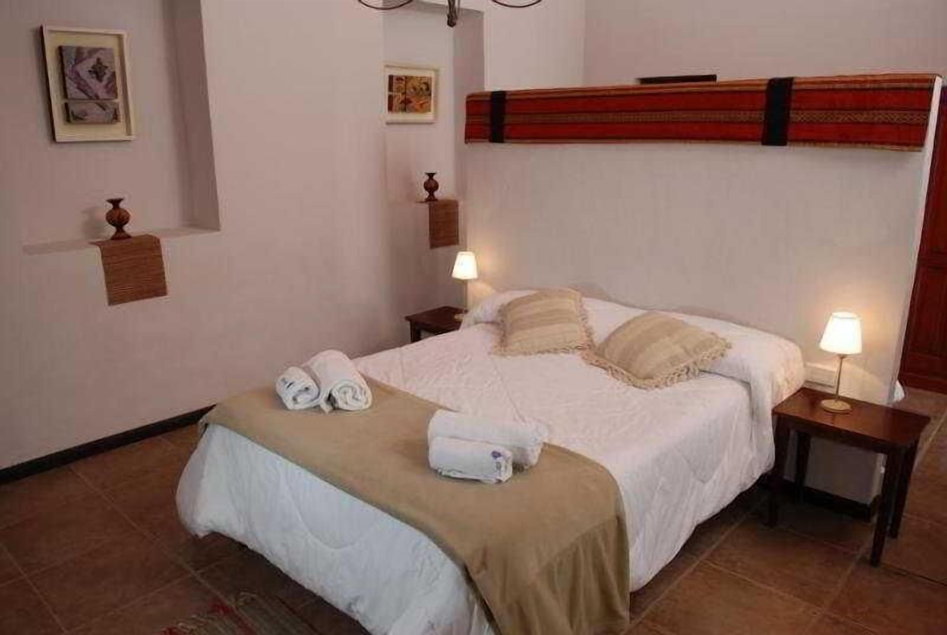 Un lit bien fait, avec du linge de lit blanc et un plaid beige, deux lampes de chevet et une petite table de nuit en bois avec une lampe.