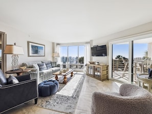 A spacious living room with a large sofa, a coffee table, and a TV mounted on the wall. There are large windows providing a view of the outdoors.