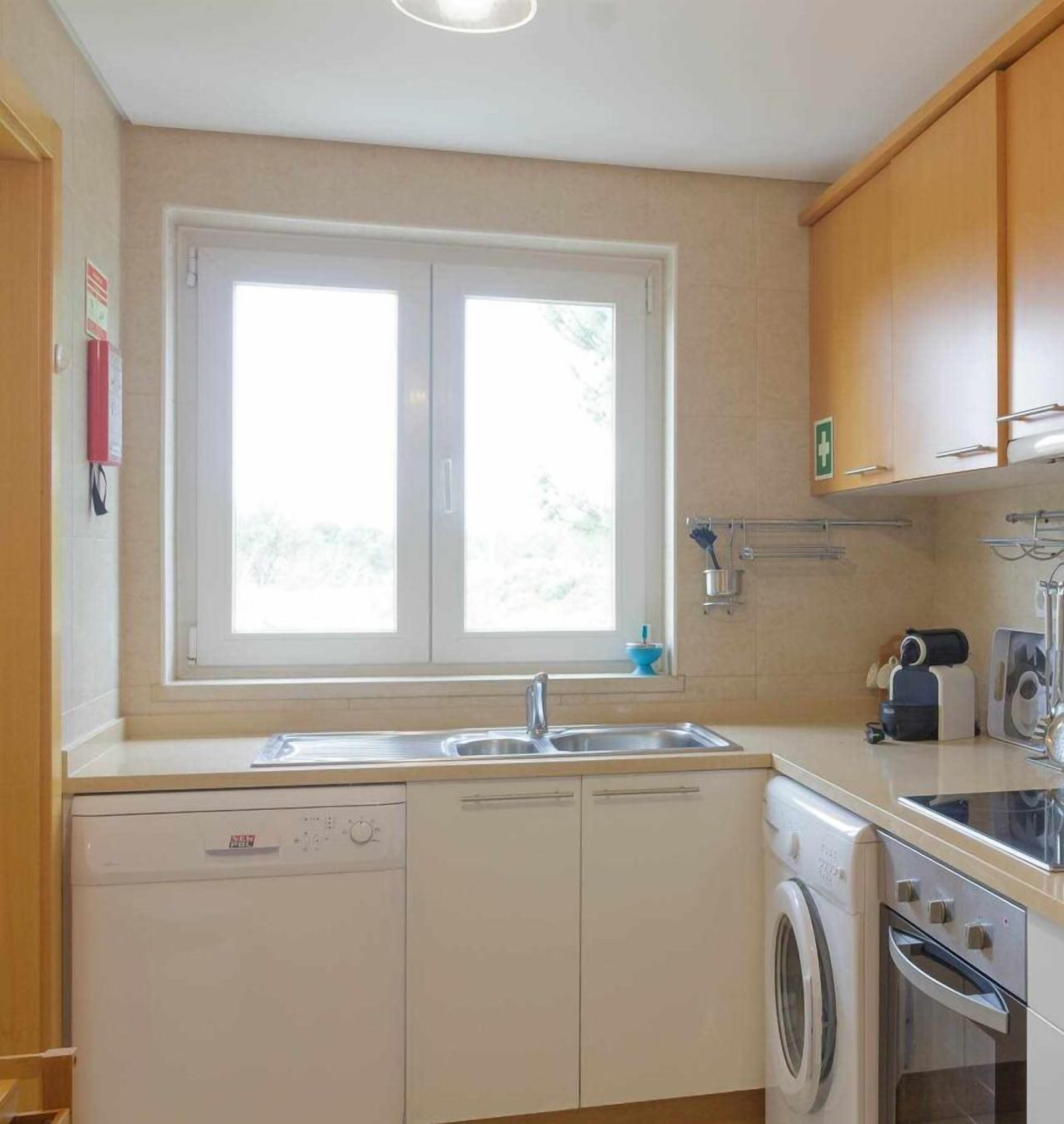 A compact kitchen with a window, a dishwasher, a washing machine, and wooden cabinets.