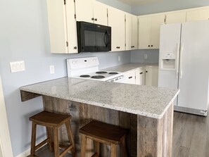 A kitchen with a granite countertop, white cabinets, a black microwave, a white stove, and a white refrigerator. There are two wooden stools at the island. - Family Getaway, Minutes away from Leland, Lake Leelanau, Suttons Bay (Lake Leelanau)