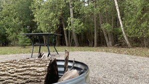 A metal fire pit with logs, surrounded by gravel, in a wooded area with a metal table and chairs. - Family Getaway, Minutes away from Leland, Lake Leelanau, Suttons Bay (Lake Leelanau)