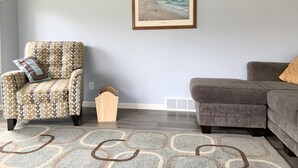 A living room with a patterned armchair, a gray sofa, a framed beach painting, and a patterned rug. - Family Getaway, Minutes away from Leland, Lake Leelanau, Suttons Bay (Lake Leelanau)