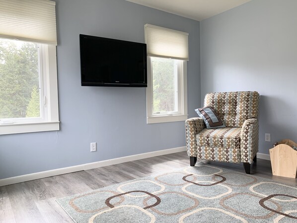 A room with a grey wall, a flat-screen TV mounted above a window, a patterned armchair, and a patterned rug. - Family Getaway, Minutes away from Leland, Lake Leelanau, Suttons Bay (Lake Leelanau)